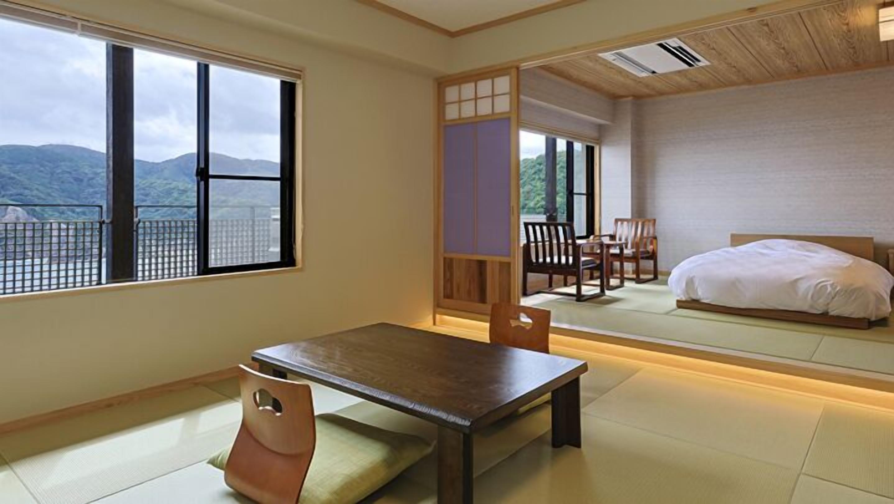 A traditional Japanese-style room with tatami flooring, a low wooden table, a cushioned chair, a bed, and large windows offering a mountain view.