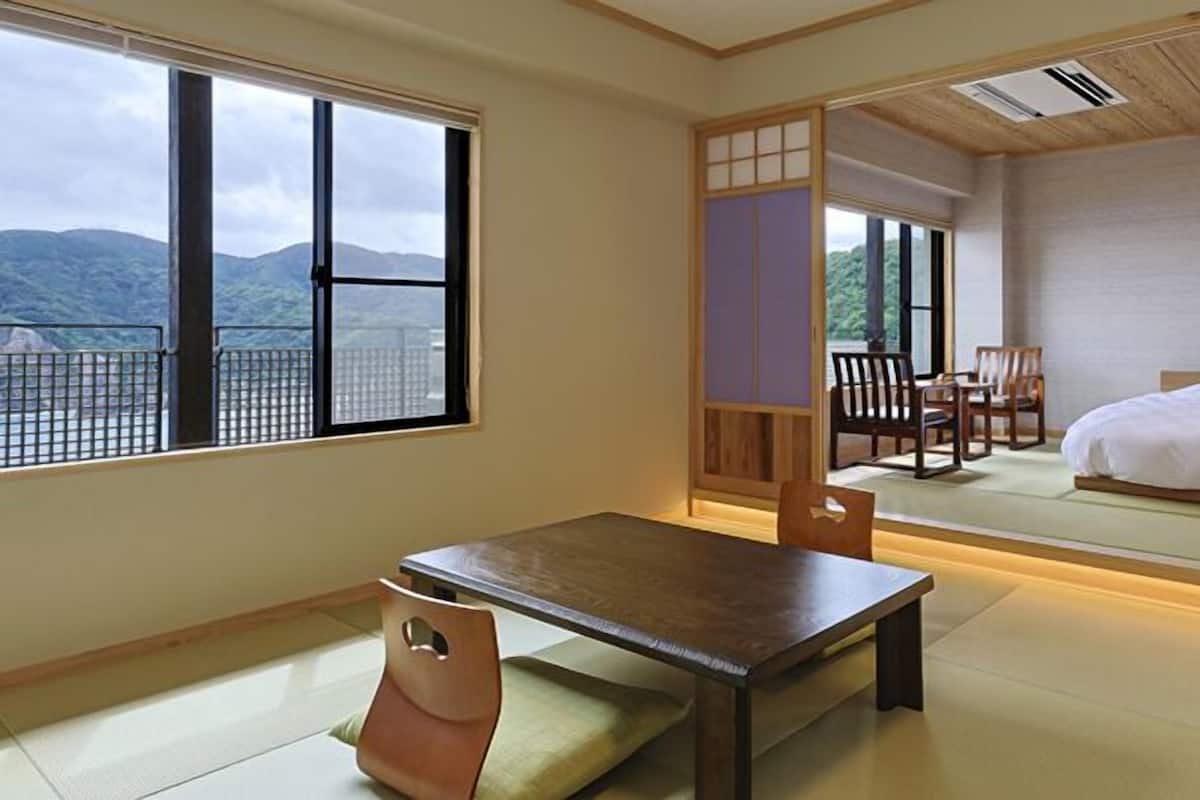 A traditional Japanese-style room with tatami flooring, a low wooden table, a cushioned chair, a bed, and large windows offering a mountain view.