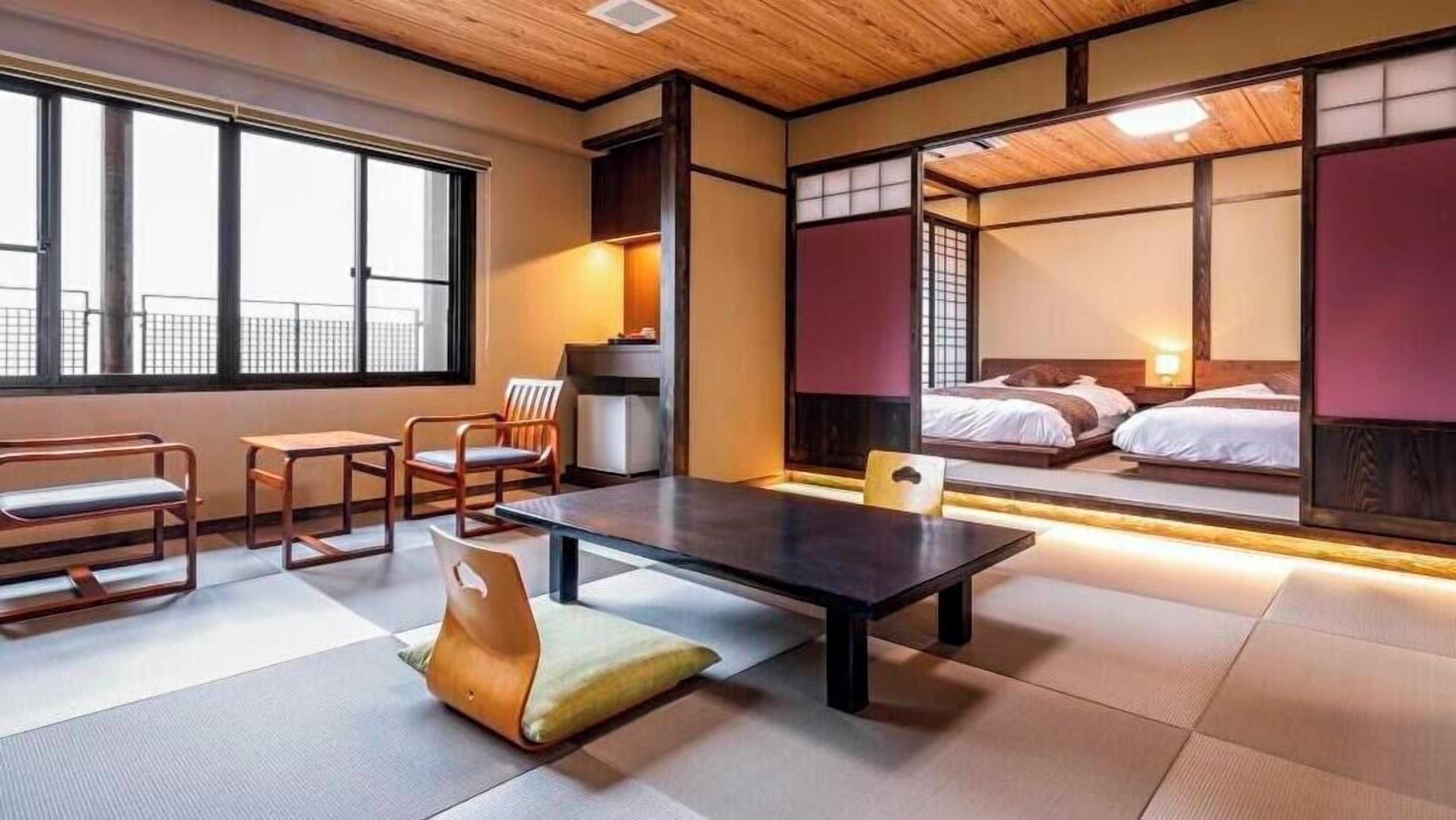 A traditional Japanese-style room with tatami flooring, a low table, chairs, and a bed with a view of the outside.