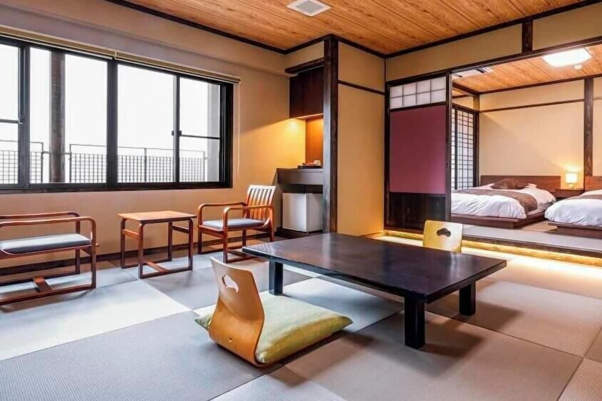 A traditional Japanese-style room with tatami flooring, a low table, chairs, and a bed with a view of the outside.