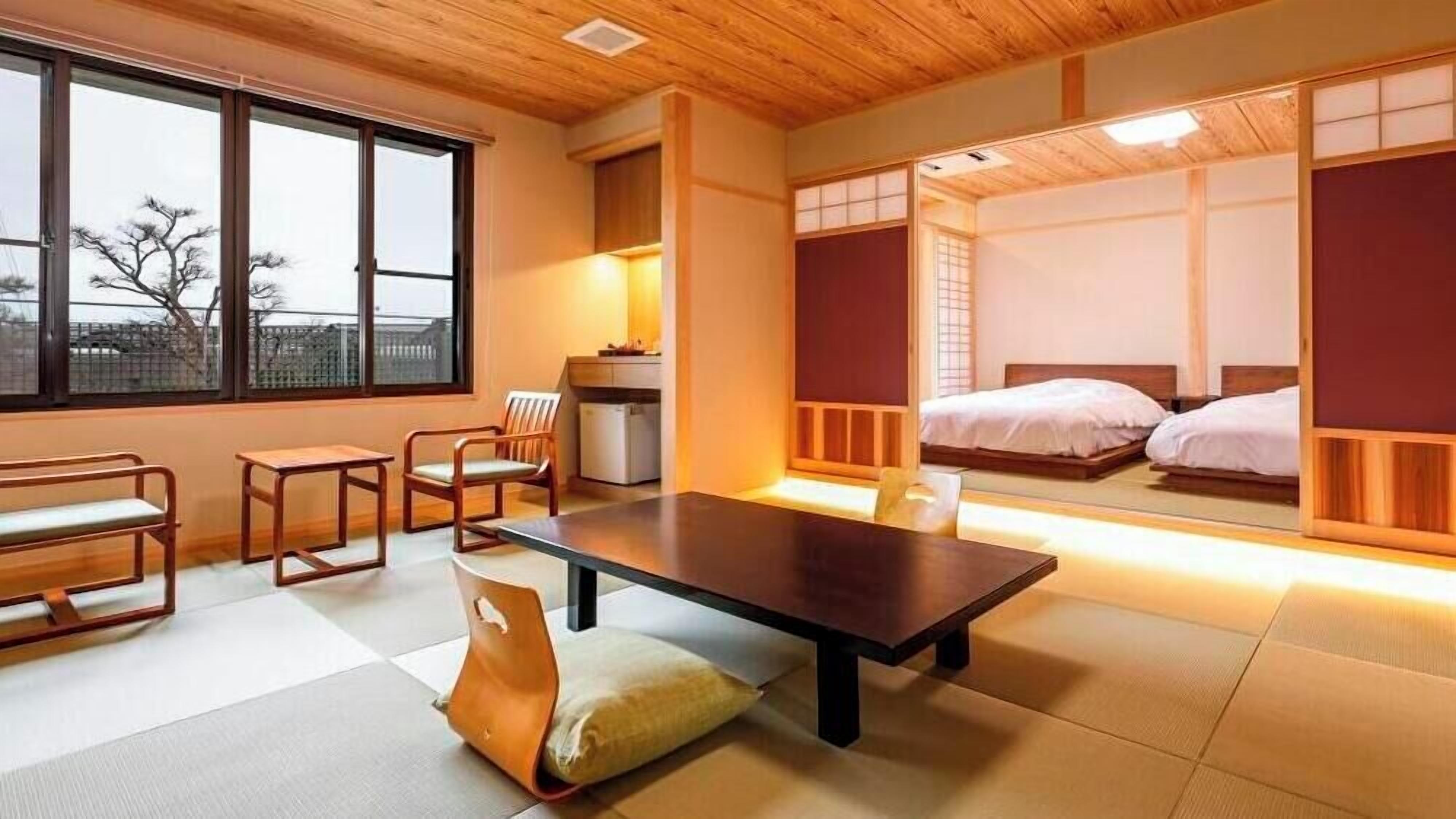 A traditional Japanese-style room with tatami flooring, a low table, a cushioned chair, a bed, and sliding doors.