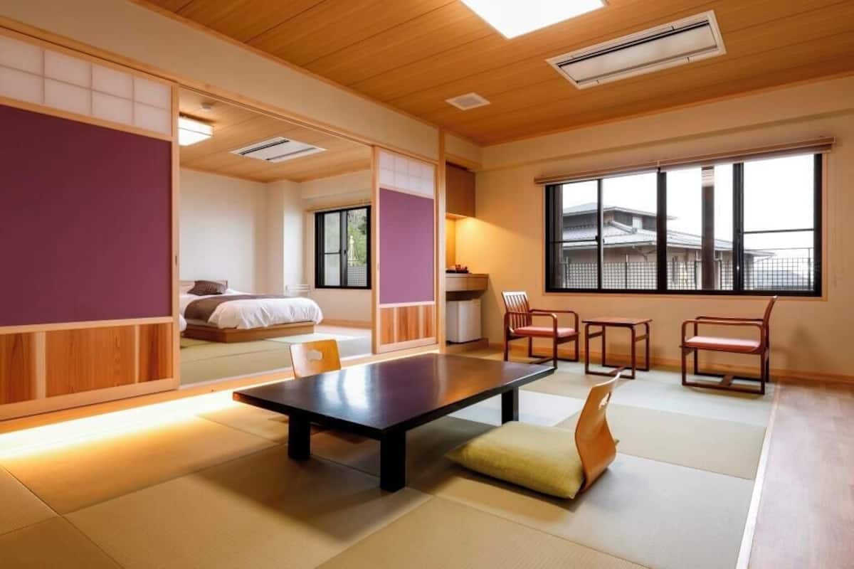 A traditional Japanese room with tatami flooring, a low table, chairs, a bed, and a large window.