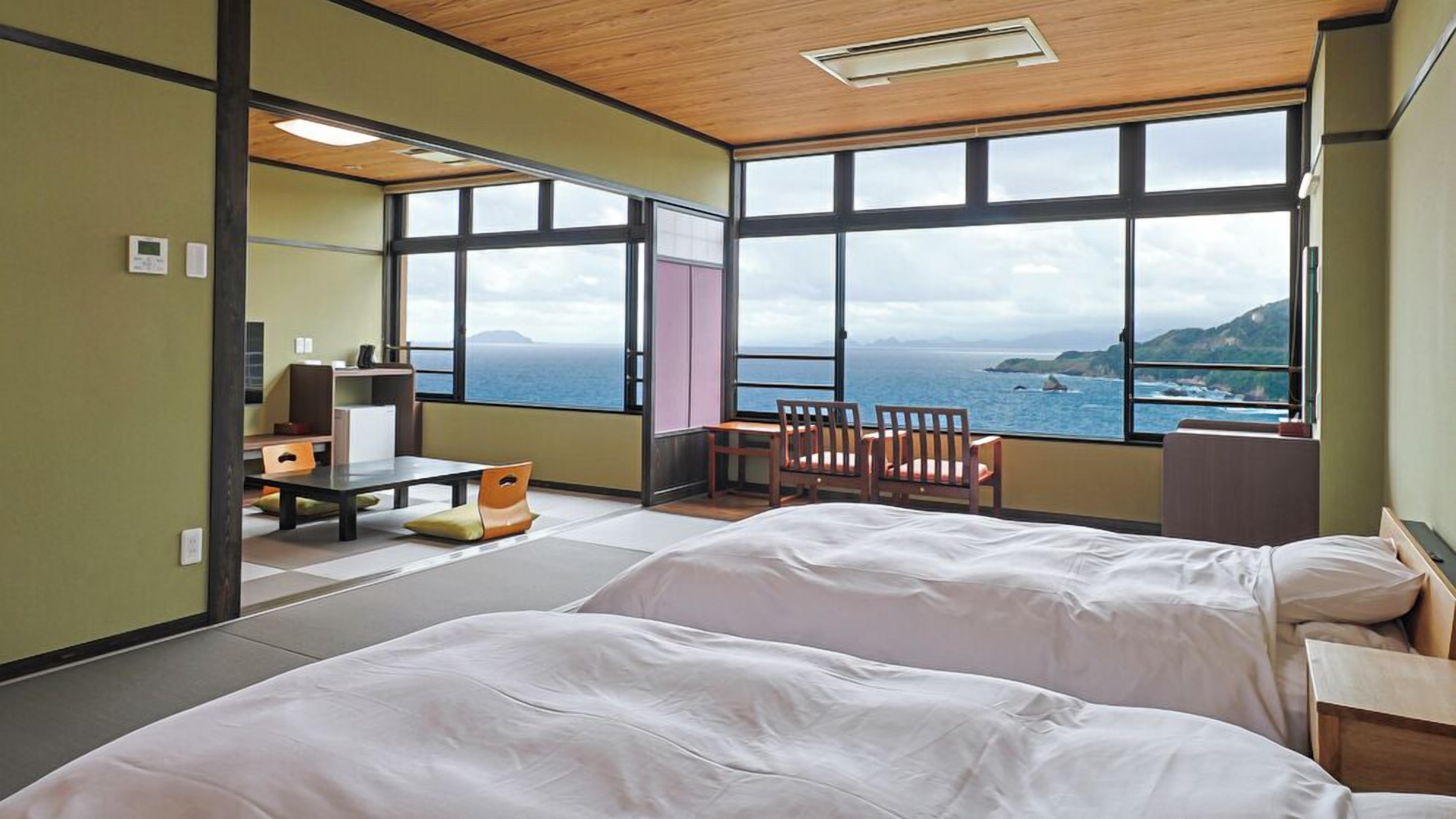 A room with two beds, a desk, a chair, and large windows offering a view of the sea.