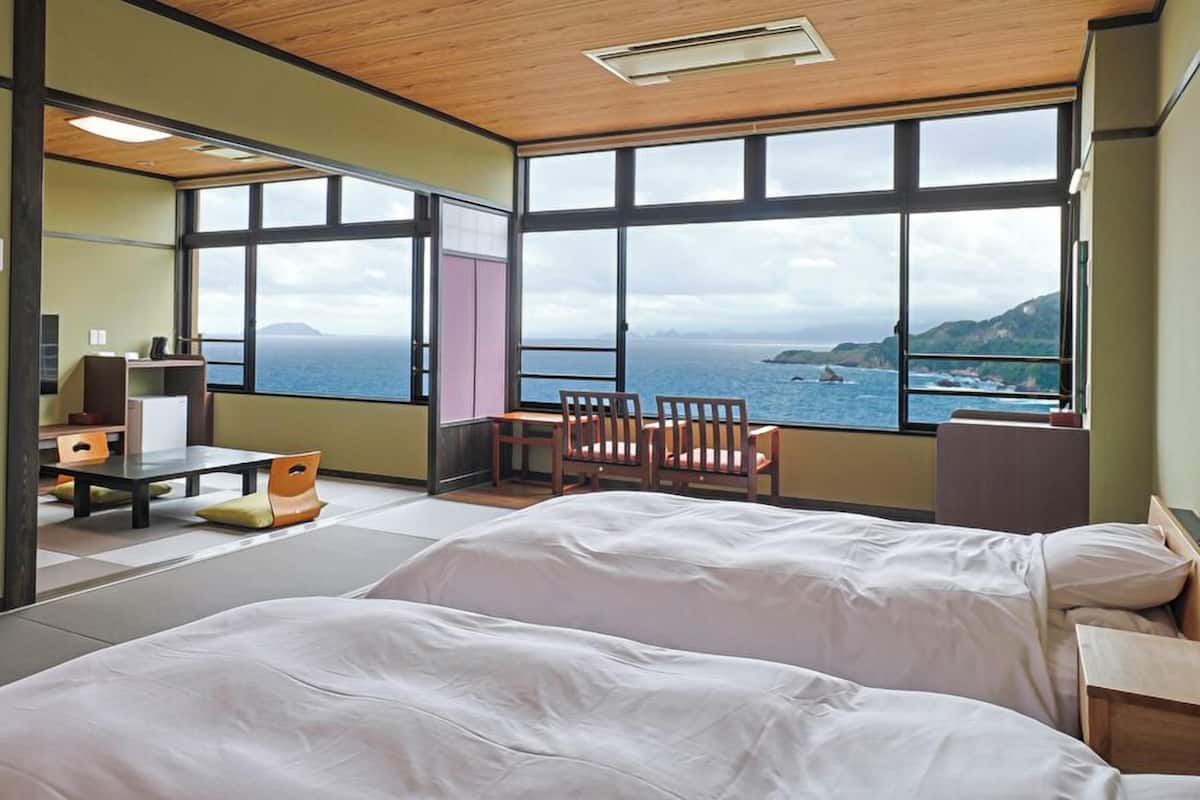 A room with two beds, a desk, a chair, and large windows offering a view of the sea.