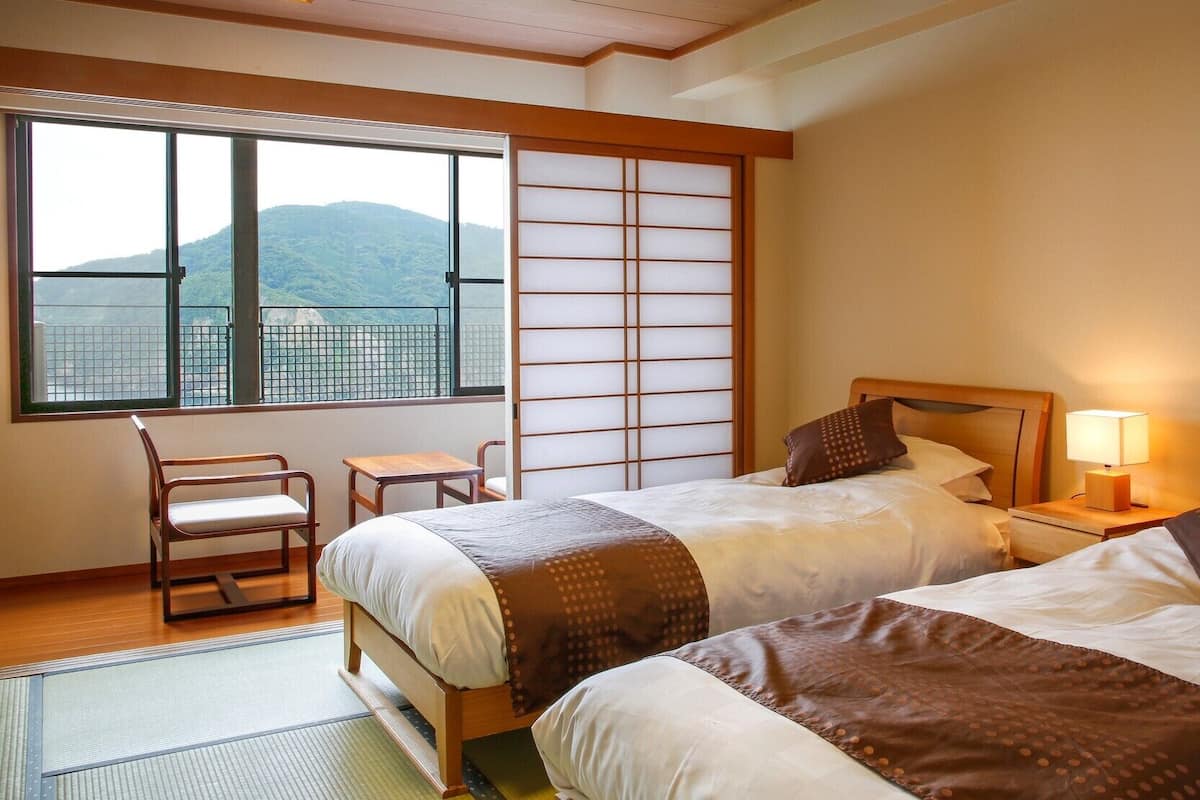 A hotel room with two beds, a chair, a small table, a window with a view of mountains, and a shoji screen.