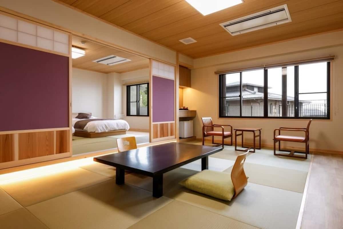 A traditional Japanese room with tatami flooring, a low table, chairs, a bed, and a large window.