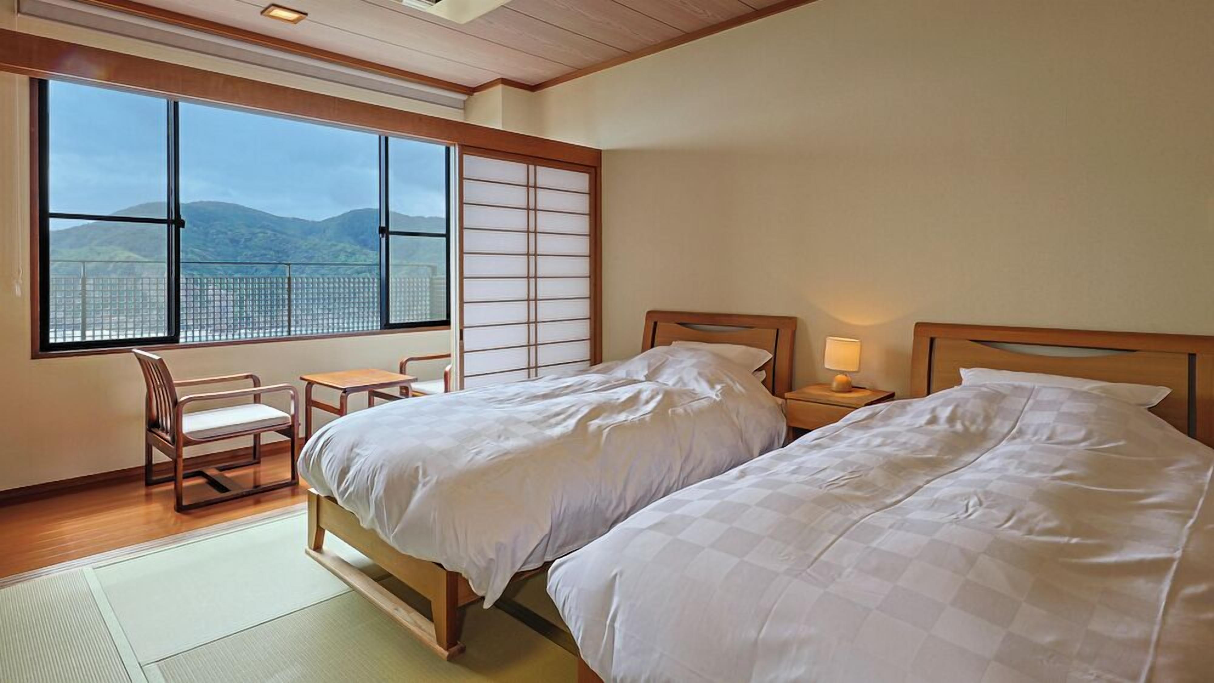 A hotel room with two beds, a desk, a chair, and a large window with a view of mountains.