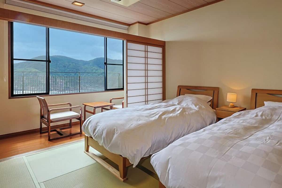 A hotel room with two beds, a desk, a chair, and a large window with a view of mountains.