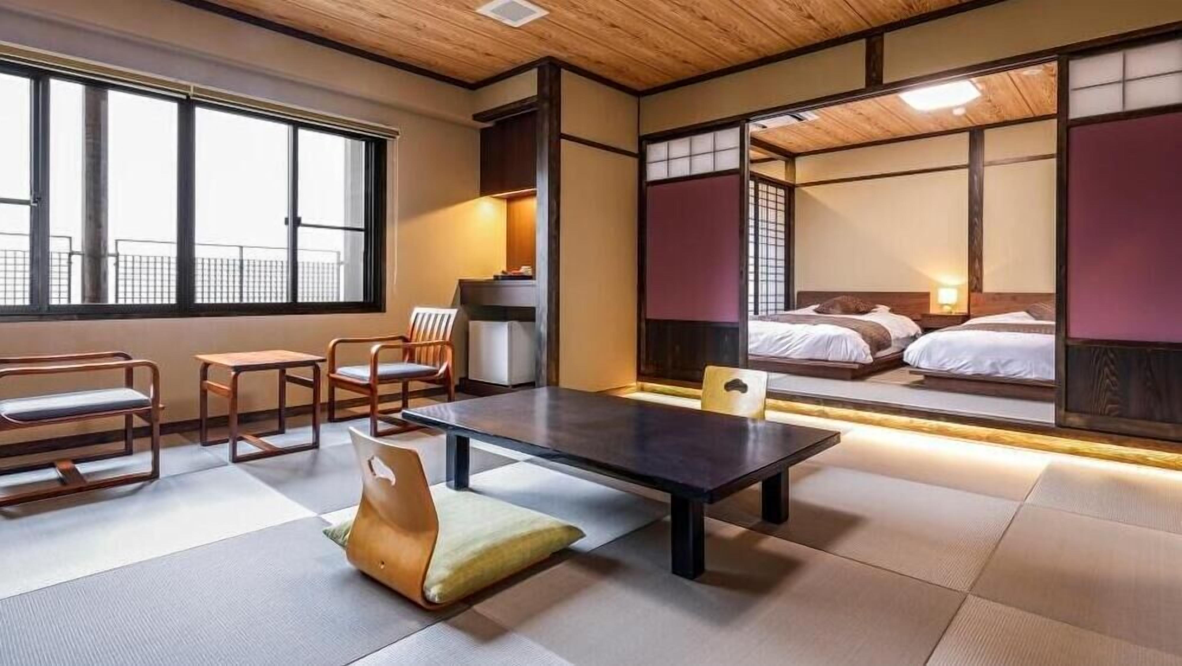 A traditional Japanese-style room with tatami flooring, a low table, chairs, and a bed with a view of the city.