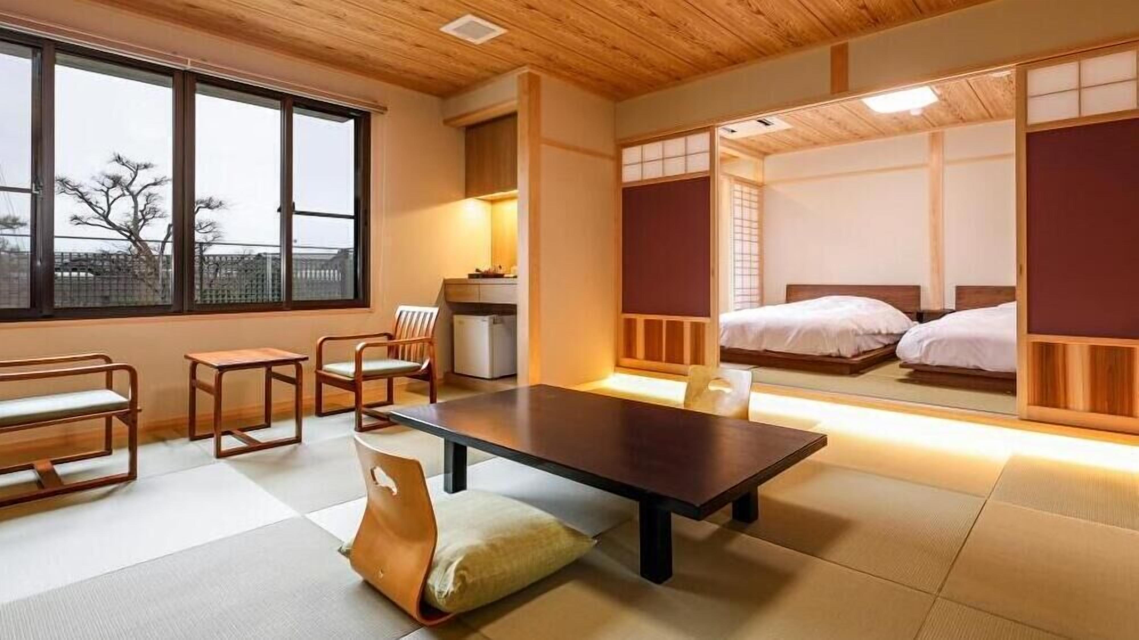 A traditional Japanese-style room with tatami flooring, a low table, a chair, and a bed with a futon.