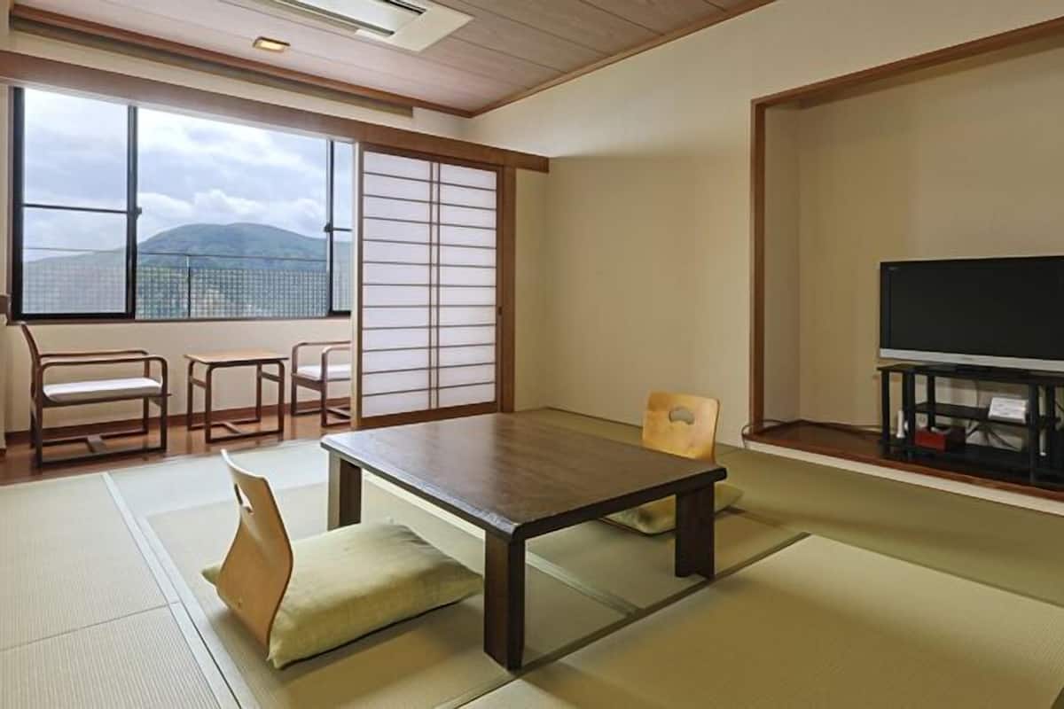 A traditional Japanese room with tatami flooring, a low table, a sofa, a TV, and a sliding door with a view of mountains.