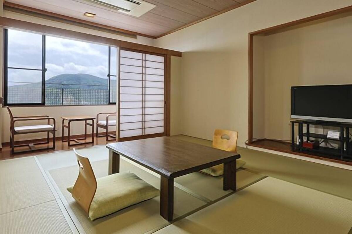 Et traditionelt japansk rum med tatamimåtter, et lavt bord, en sofa, et fjernsyn og en skydedør med udsigt til bjerge.