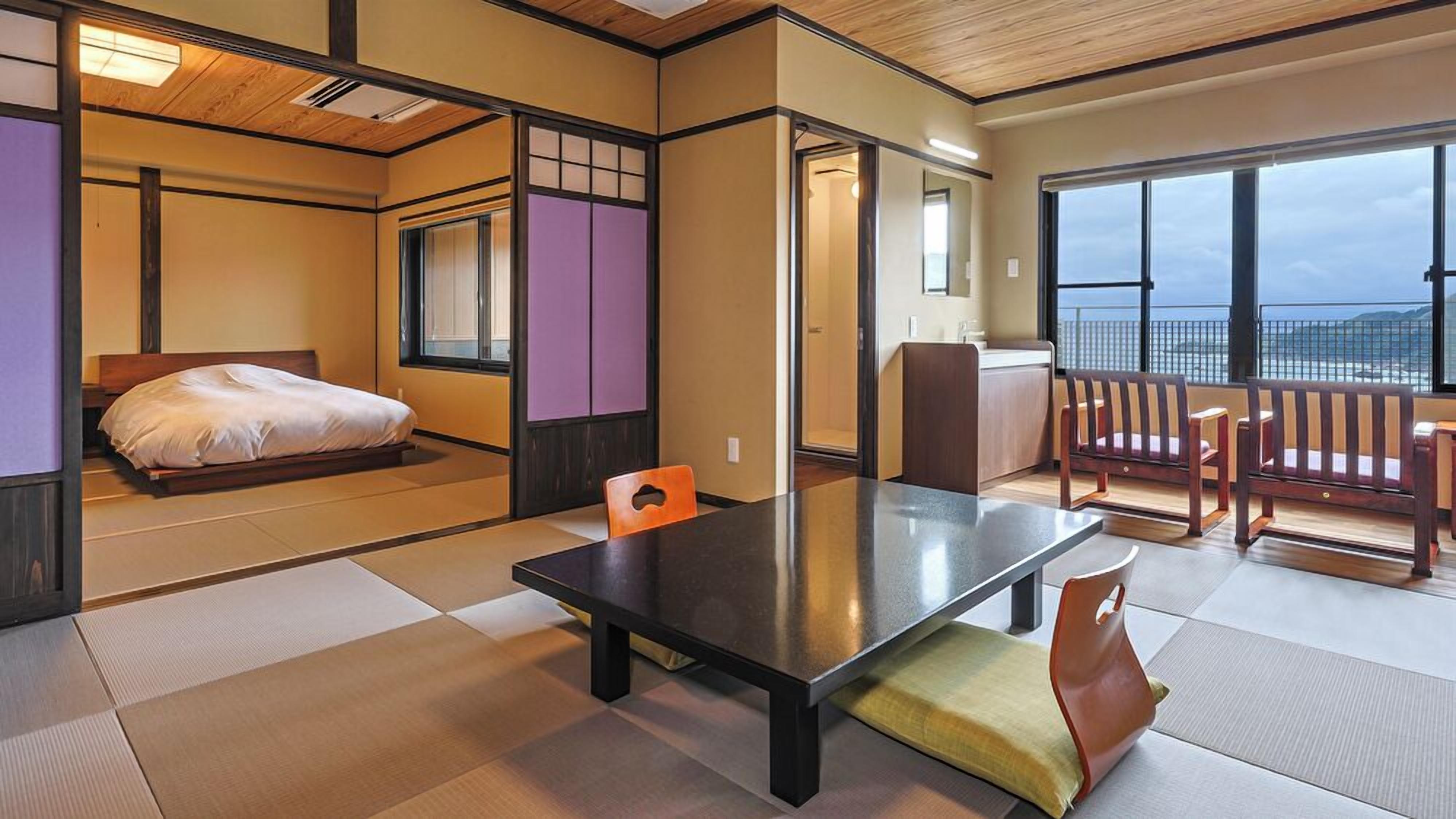 A traditional Japanese room with tatami flooring, a bed, a table, and a view of the sea.