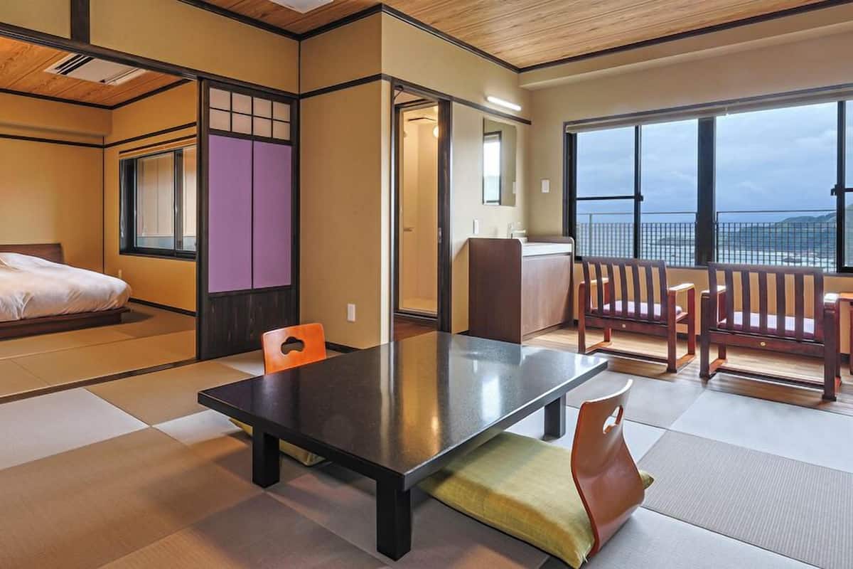 A traditional Japanese room with tatami flooring, a bed, a table, and a view of the sea.