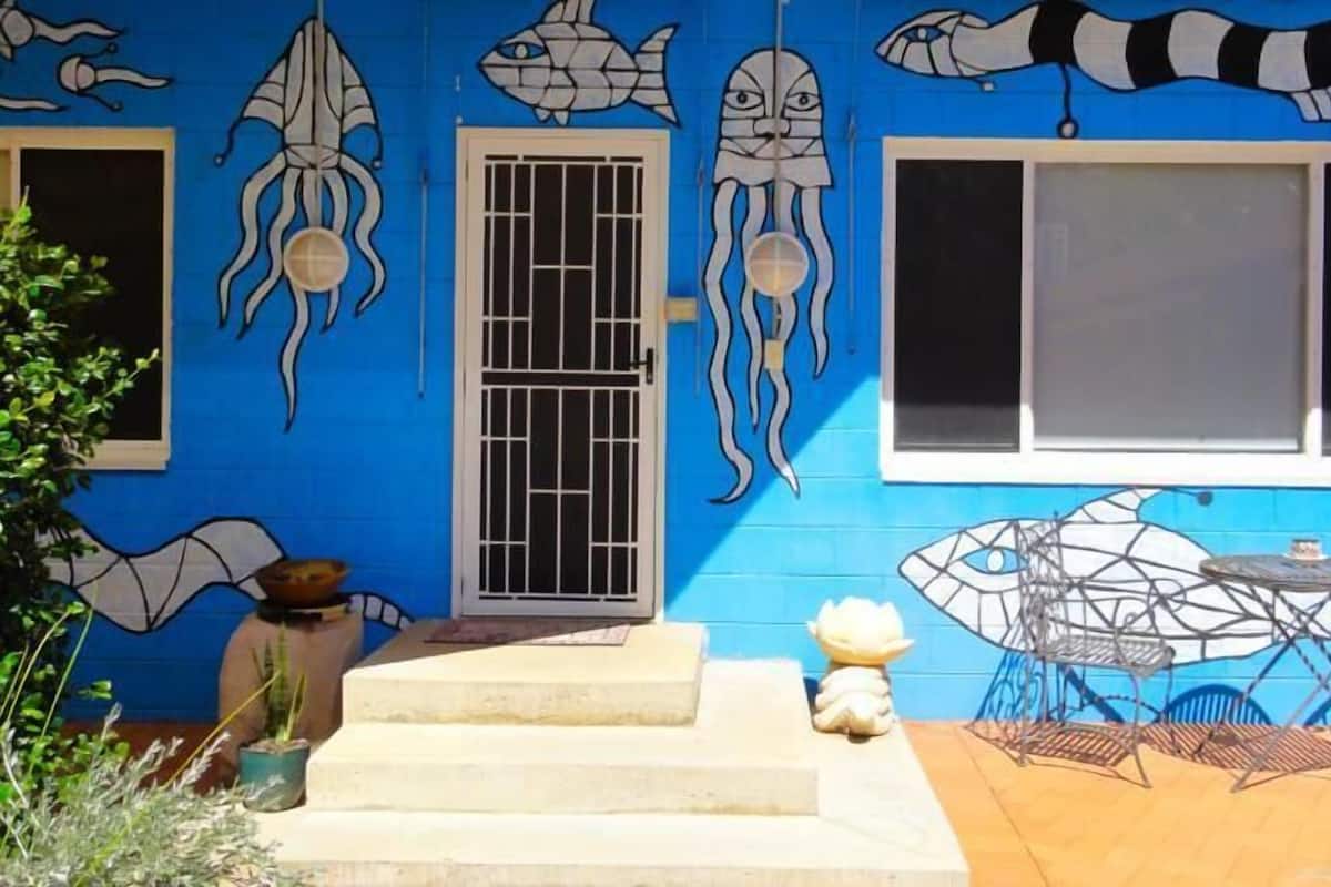 A building with a blue exterior, a white door, and a window with a metal grill. There are steps leading to the door, and a small table with chairs outside.