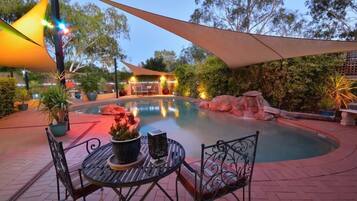 Un espace piscine aménagé avec une table et des chaises, entouré de plantes en pot et surmonté d’un grand parasol.