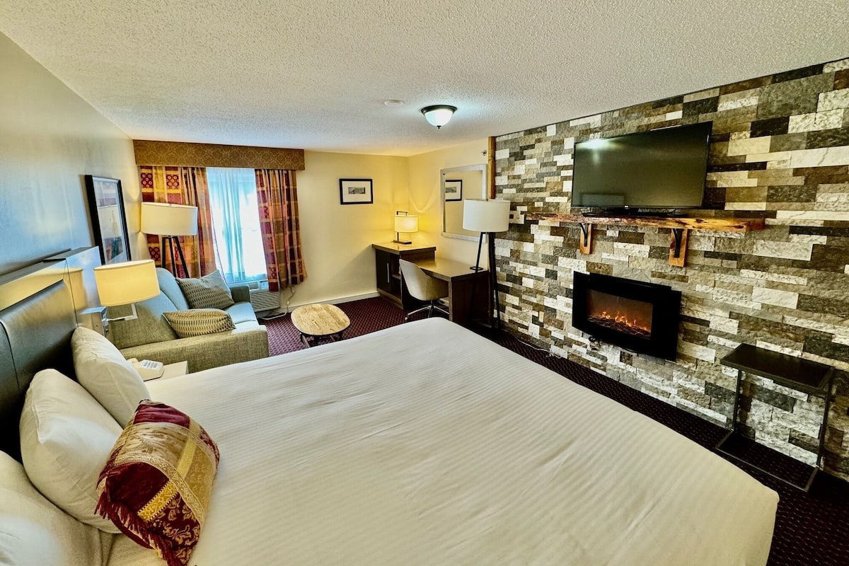 A hotel room with a large bed, a desk, a TV mounted on a stone wall, and a fireplace.