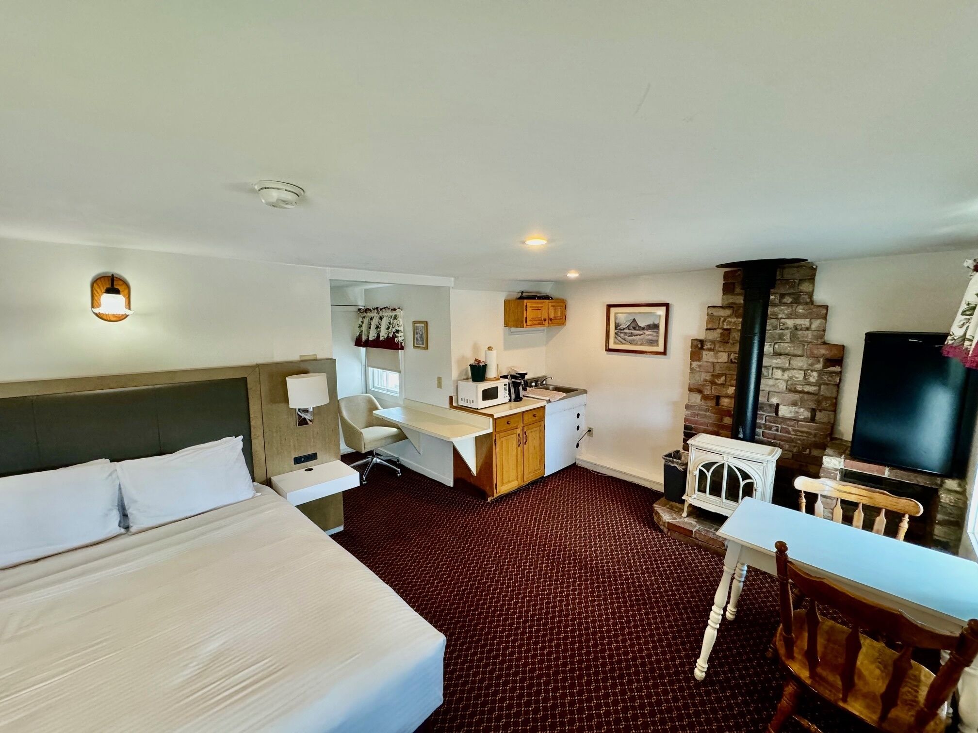 A hotel room with a bed, a desk, a chair, a microwave, a small kitchen area, a TV, and a fireplace.