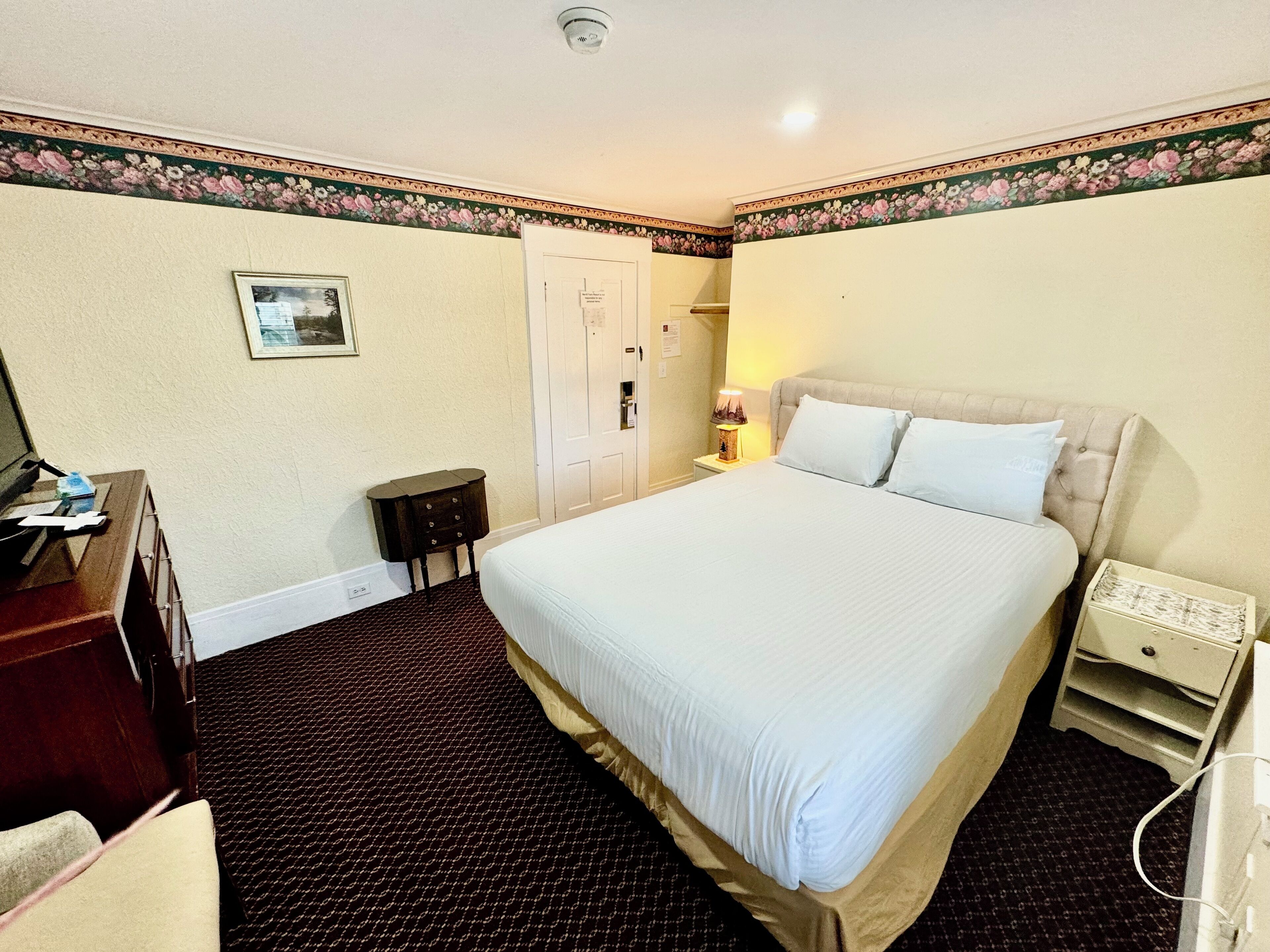 A hotel room with a large bed, a desk, a chair, a nightstand with a lamp, and a door.