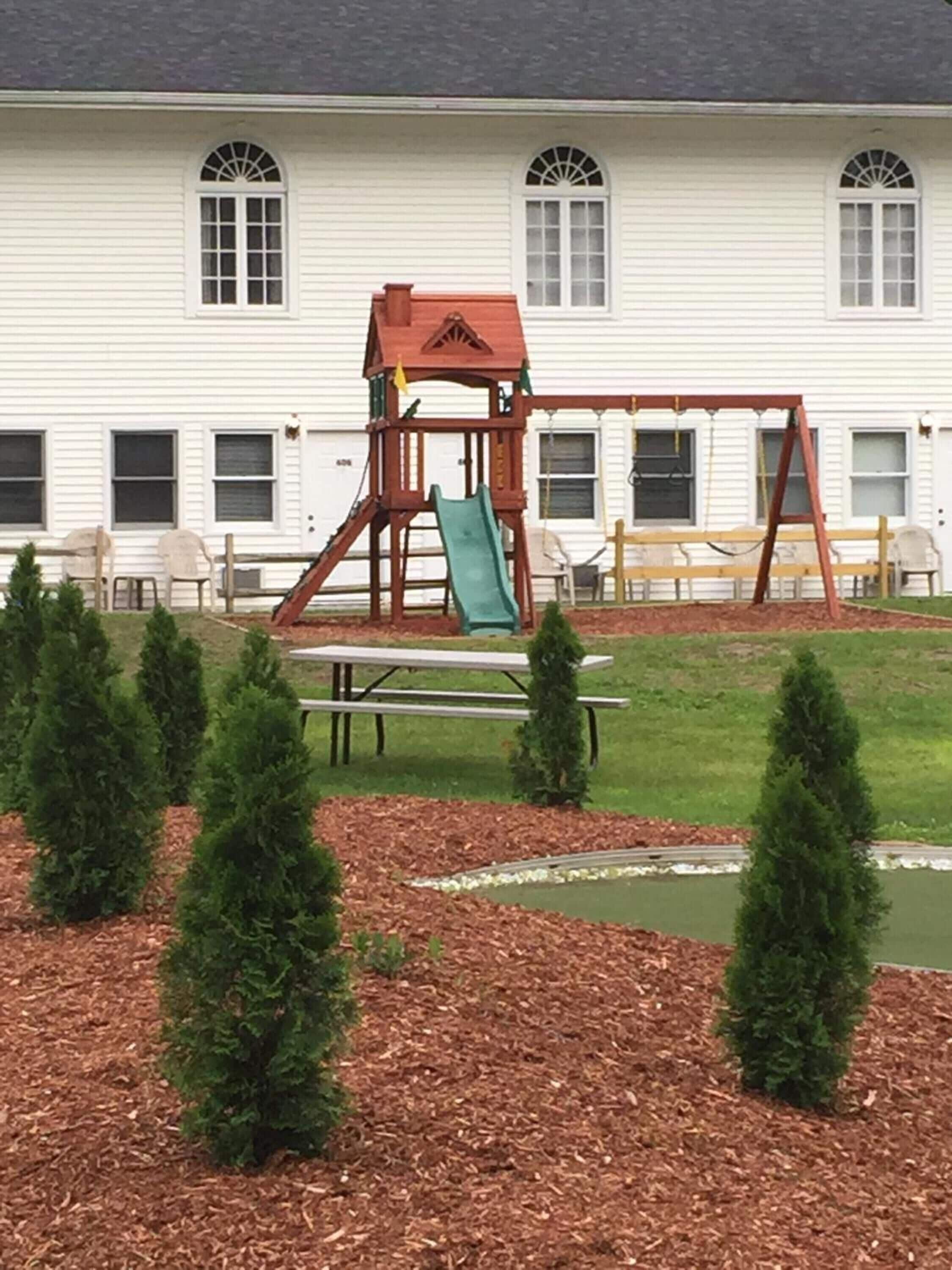 A playground with a swing set and slide, surrounded by a lawn and small trees.