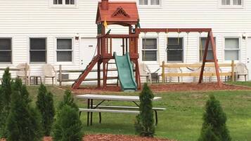 A playground with a swing set and slide, surrounded by a lawn and small trees.