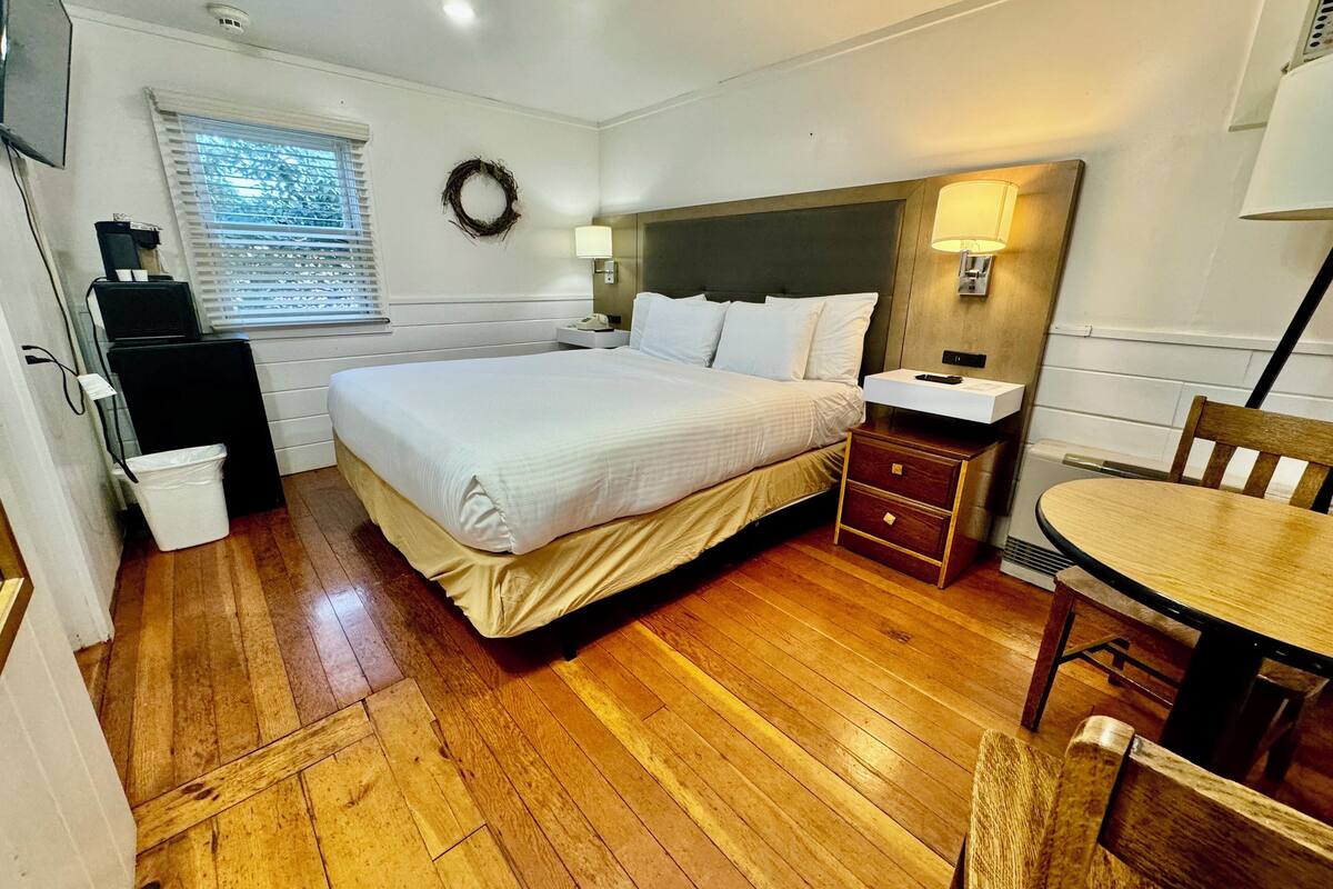 A hotel room with a large bed, bedside tables, a desk with a chair, a window with blinds, and a wall-mounted lamp.