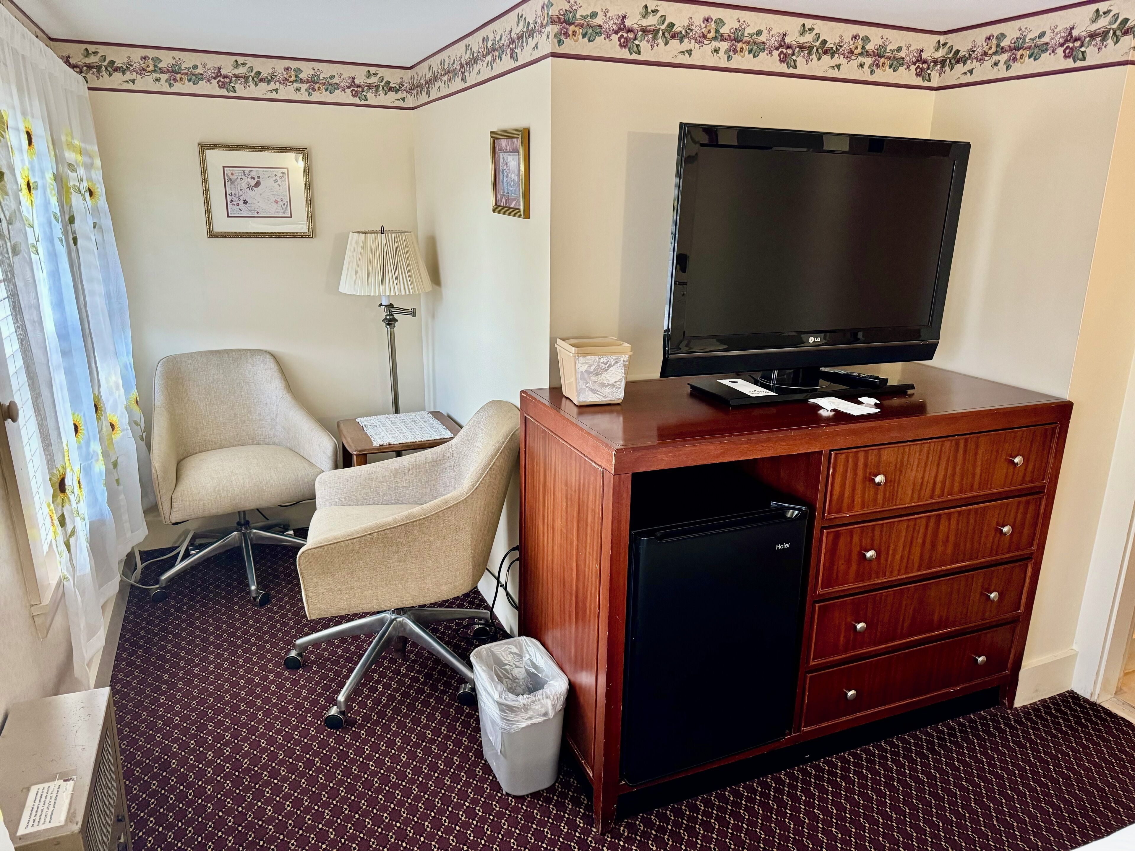 A hotel room with a wooden desk, a television, a chair, a lamp, and a trash bin.