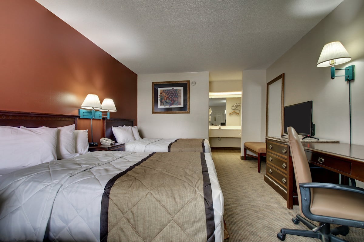 A hotel room with two beds, a desk, a chair, a dresser, a lamp, a painting, and a mirror.