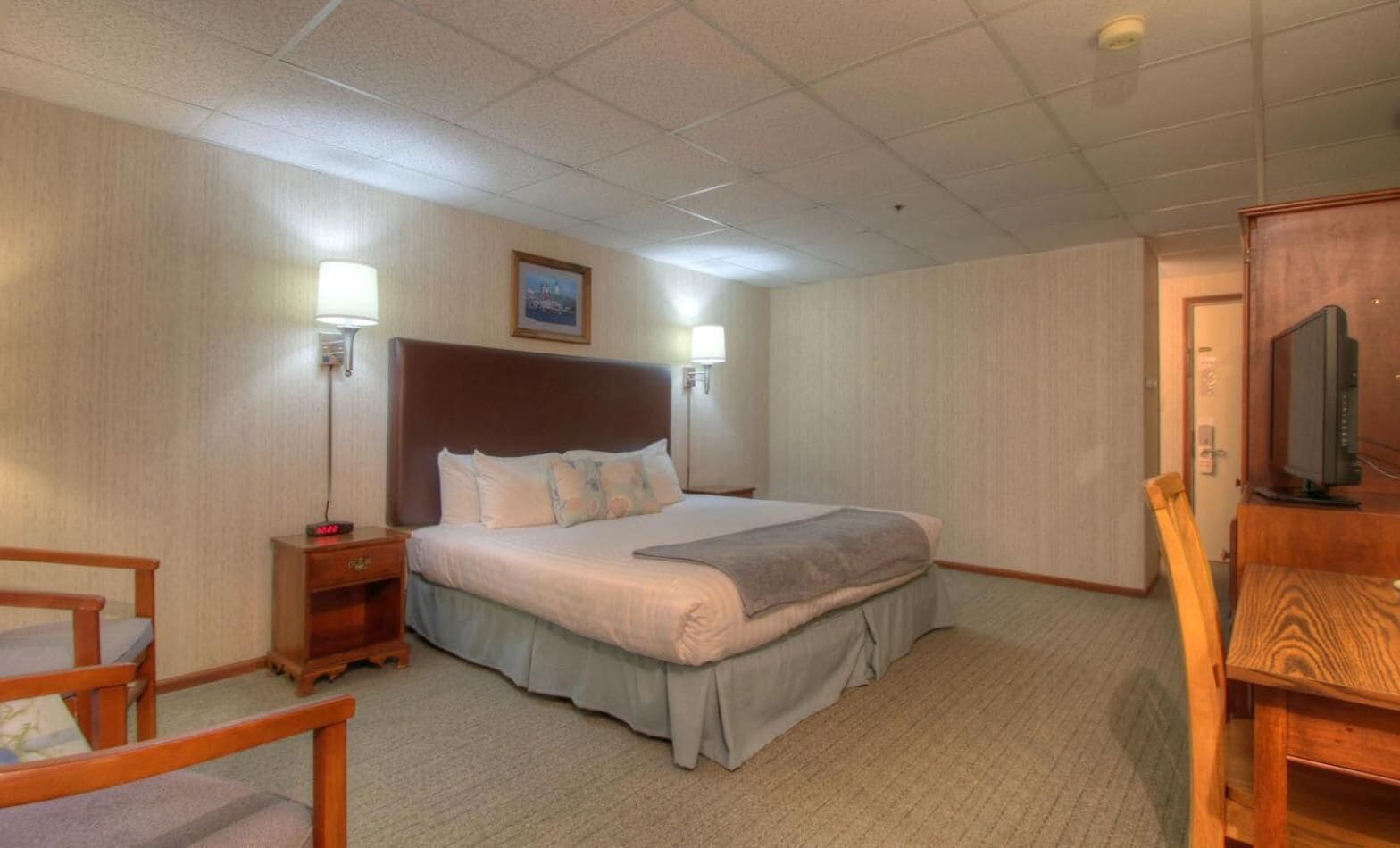 Een hotelkamer met een groot bed, een nachtkastje met een lamp, een stoel, een bureau met een televisie en een schilderij aan de muur.
