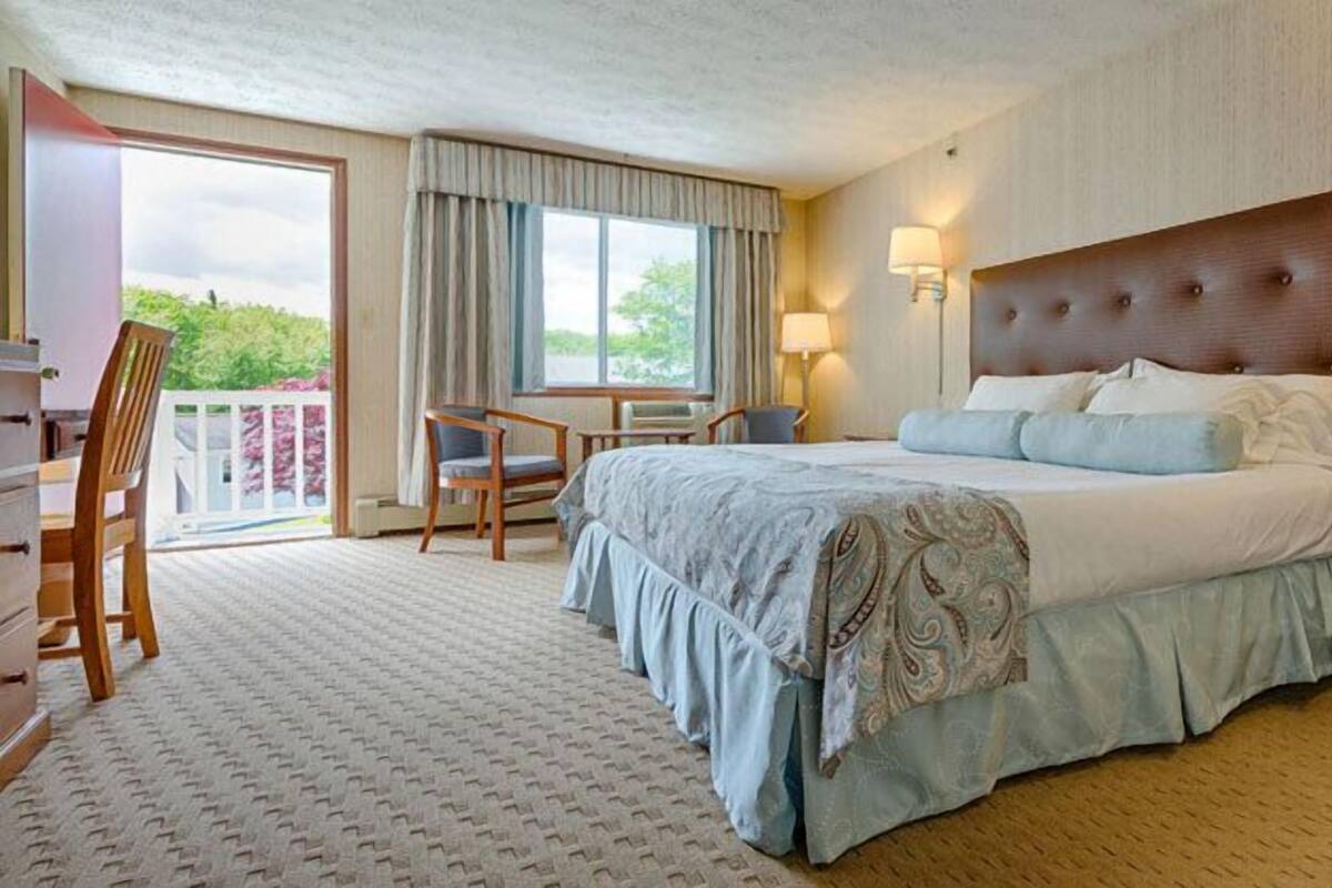 A hotel room with a large bed, a desk with a chair, a balcony with a view, and a nightstand with a lamp.