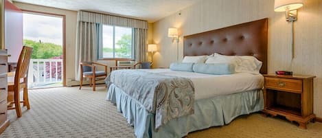 A hotel room with a large bed, a desk with a chair, a balcony with a view, and a nightstand with a lamp.