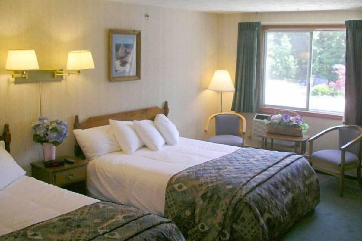 A hotel room with two beds, a desk, two lamps, a chair, and a window with curtains.