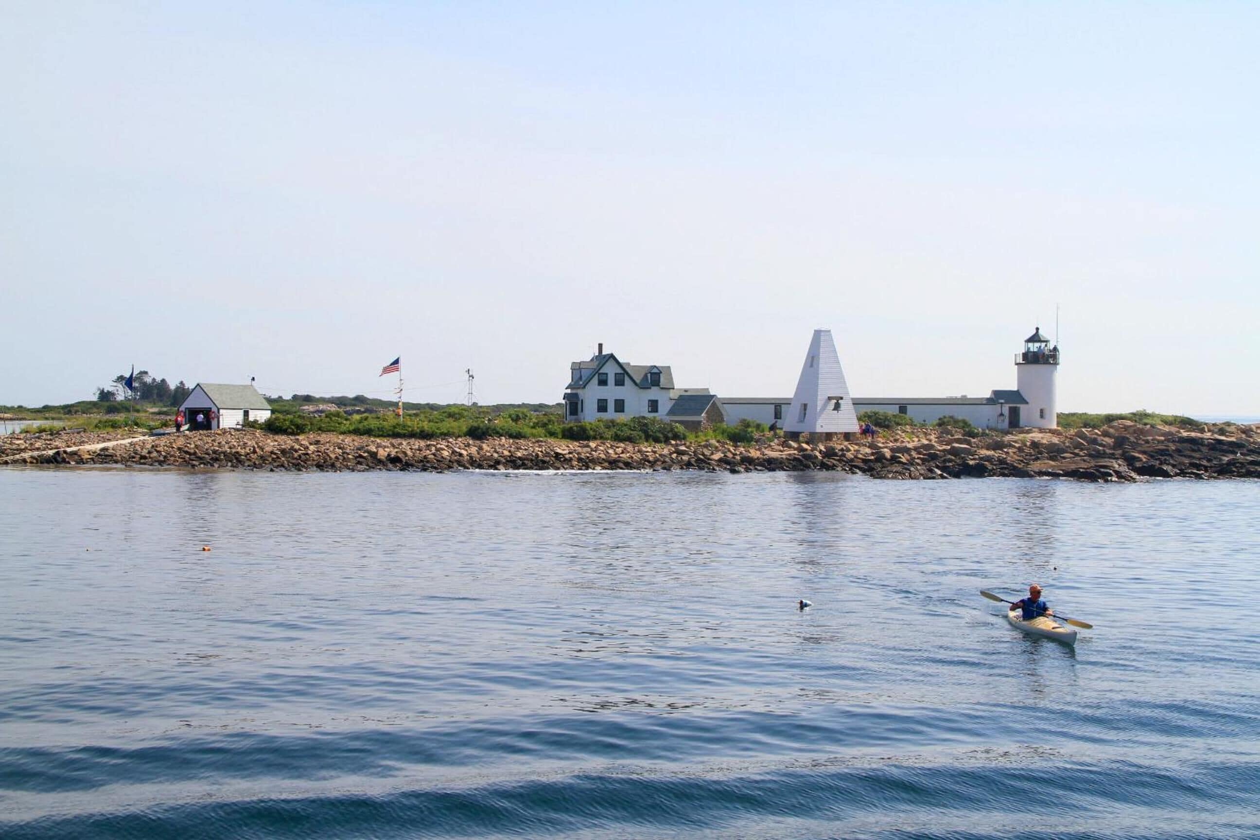 Un paisaje costero con un faro, un edificio blanco con techo puntiagudo y una orilla rocosa.