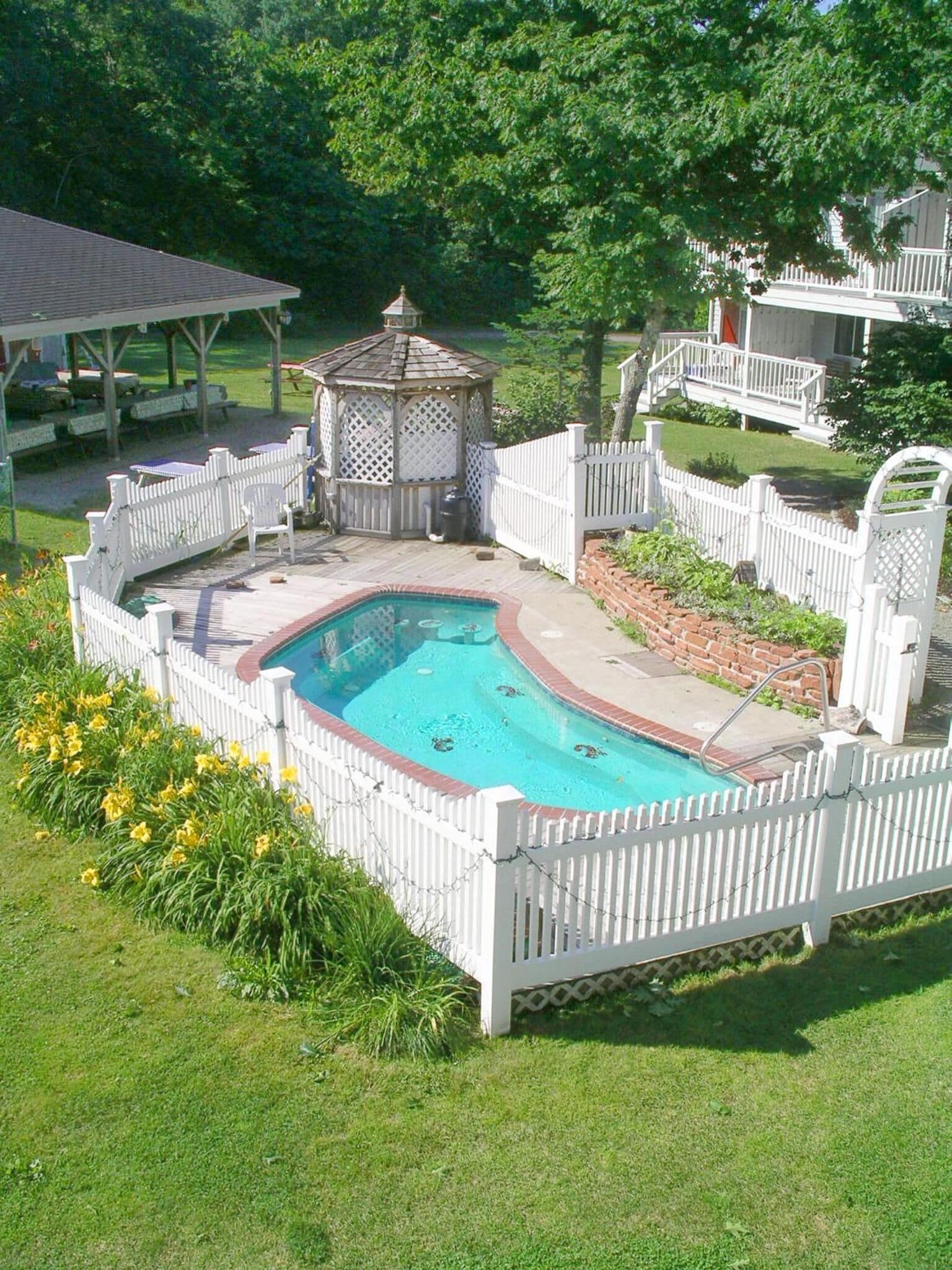 A backyard with a swimming pool surrounded by a white picket fence, a gazebo, and a covered patio area.