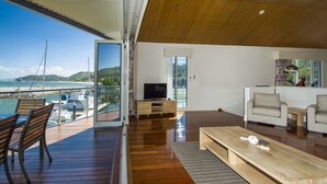 A modern living room with a wooden floor, a large window, and a view of the sea. - Waters Edge Townhouse 4 (Nelly Bay)