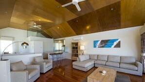 A spacious living area with a wooden ceiling, a large sofa, a coffee table, and a kitchen in the background. - Waters Edge Townhouse 4 (Nelly Bay)