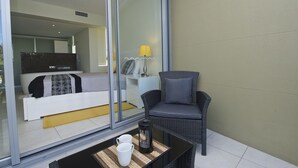 Un balcón con una silla de mimbre, una mesita con dos tazas blancas, una cafetera de vidrio y una cama con ropa de cama estampada.