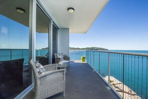 Uma varanda com móveis de vime, guarda-corpo de vidro e vista para o mar.