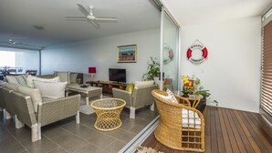 A modern living room with wicker furniture, a ceiling fan, a TV, and a glass door leading to an outdoor area. - 1 Bright Point Apartment 5404 (Nelly Bay)