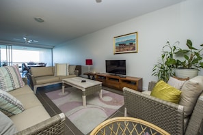 A living room with a sofa, coffee table, TV, and a painting on the wall. - 1 Bright Point Apartment 5404 (Nelly Bay)