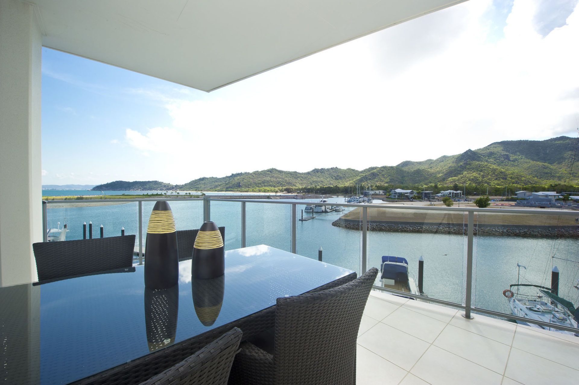 A balcony with a glass railing overlooking a marina with boats and hills in the background.