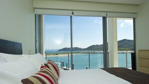 A hotel room with a large bed, a view of a beach and water, and a balcony with a railing. - Blue On Blue Superior Studio Room 1361 (Nelly Bay)