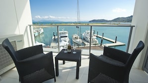 Een balkon met twee rieten stoelen en een kleine tafel, met uitzicht op een jachthaven met verschillende boten die aanmeren.