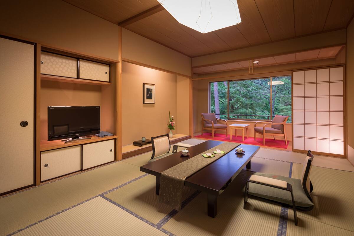 Ett traditionellt japanskt rum med tatamimattor på golvet, ett lågt bord med en löpare, en tv på ett skåp och skjutdörrar som leder ut till en balkong med utsikt över träd.