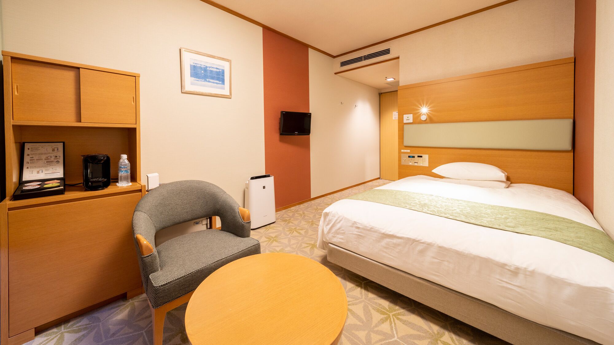 A hotel room with a bed, a chair, a small table, a TV, a heater, and a shelf with a framed picture and a coffee maker.