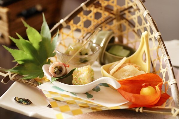 A wicker basket containing various food items, including a bowl of green sauce, a small dish with a yellow item, and a plate with a rolled pastry. - Hanamaki Onsen Kashoen (Hanamaki)