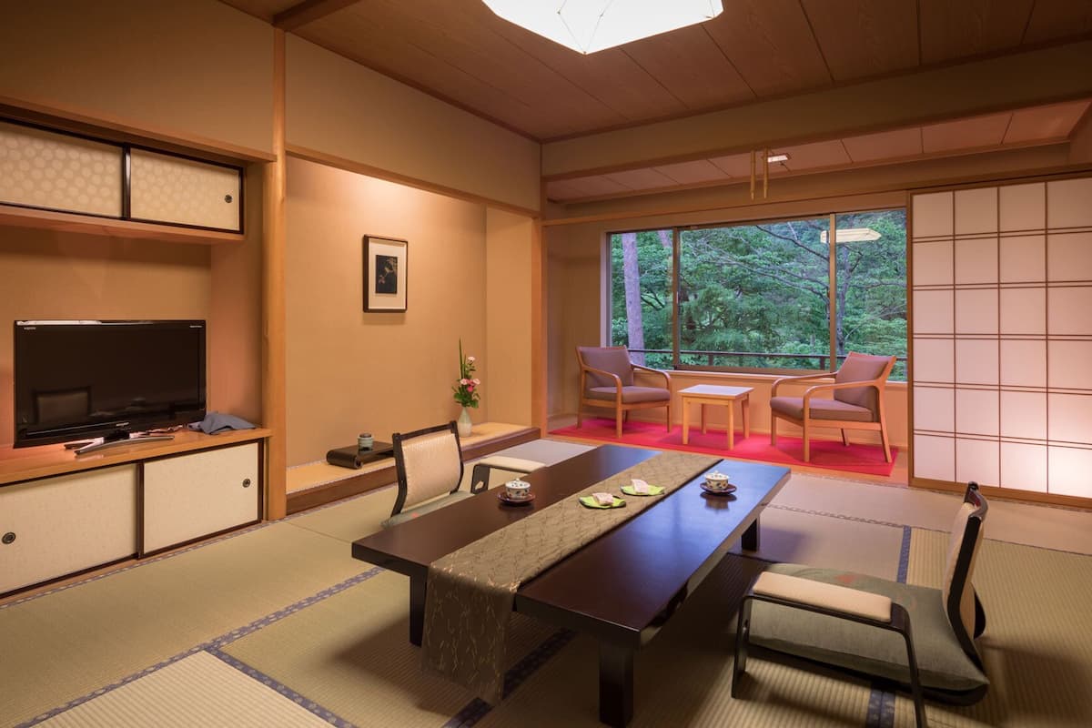 Ett traditionellt japanskt rum med tatamimattor på golvet, ett lågt bord, stolar, en tv och en skjutdörr som leder ut till en balkong med utsikt över träd.