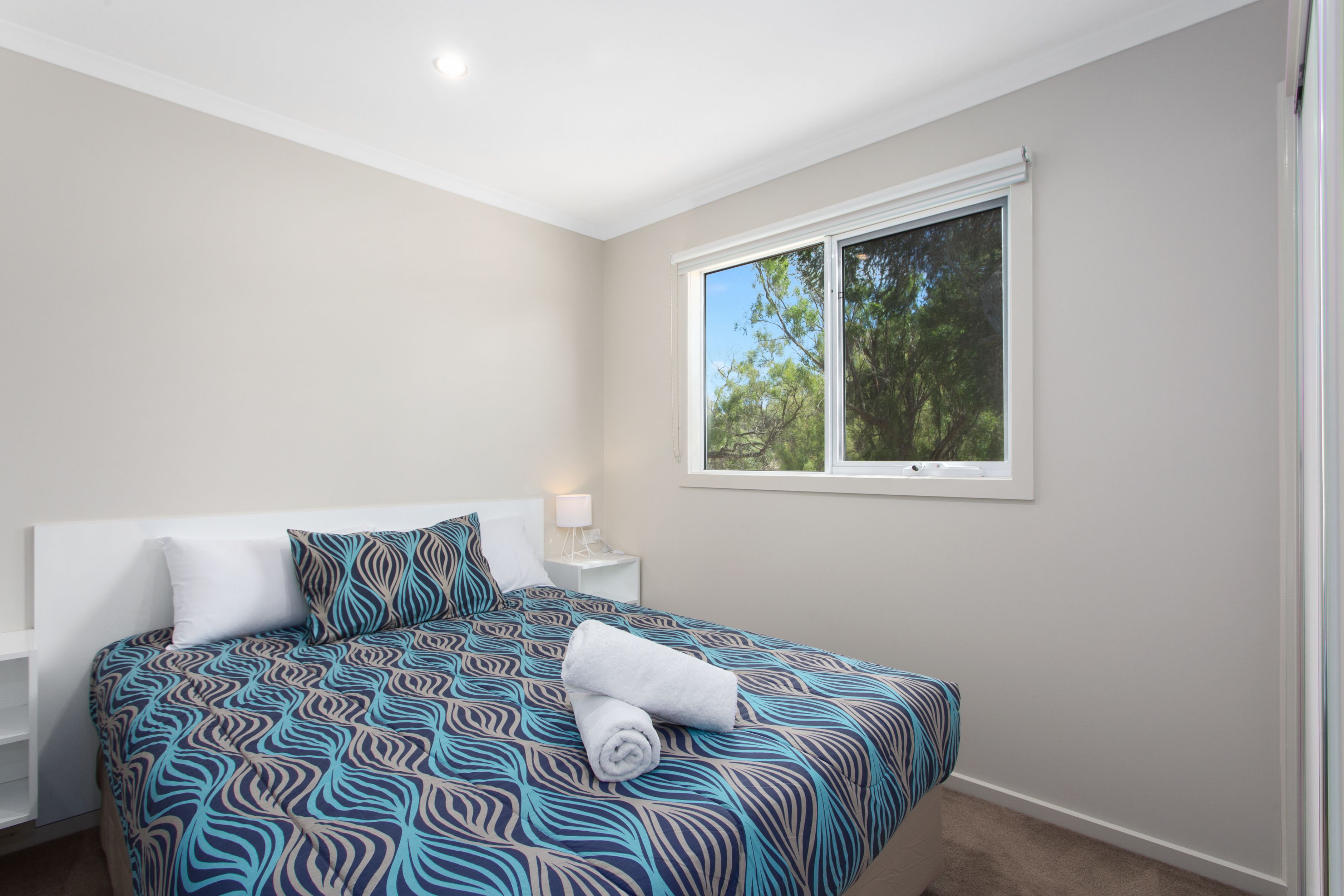 A bedroom with a bed, a nightstand, a window, and a view of trees.