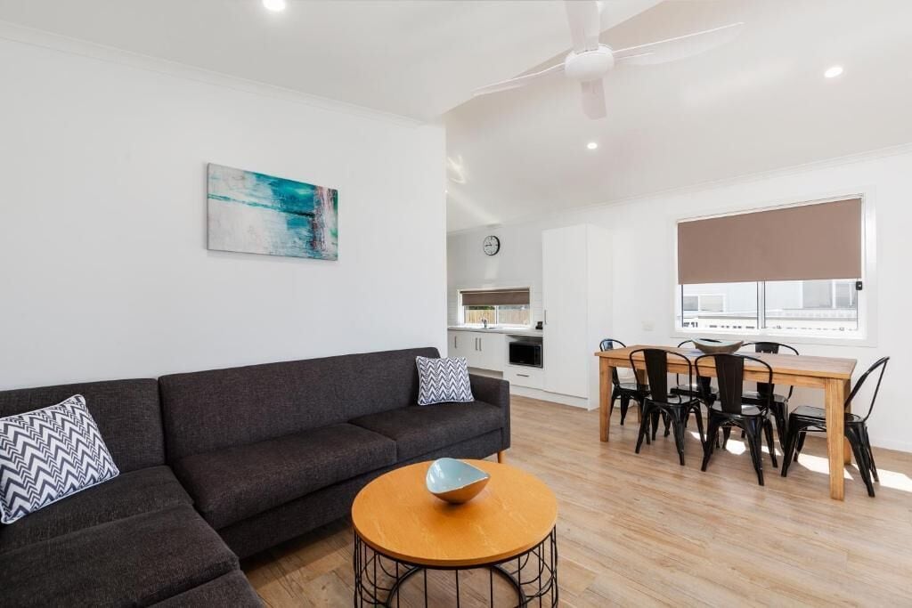 A modern living room with a dark grey sofa, a round wooden coffee table, and a dining area with a wooden table and black chairs.