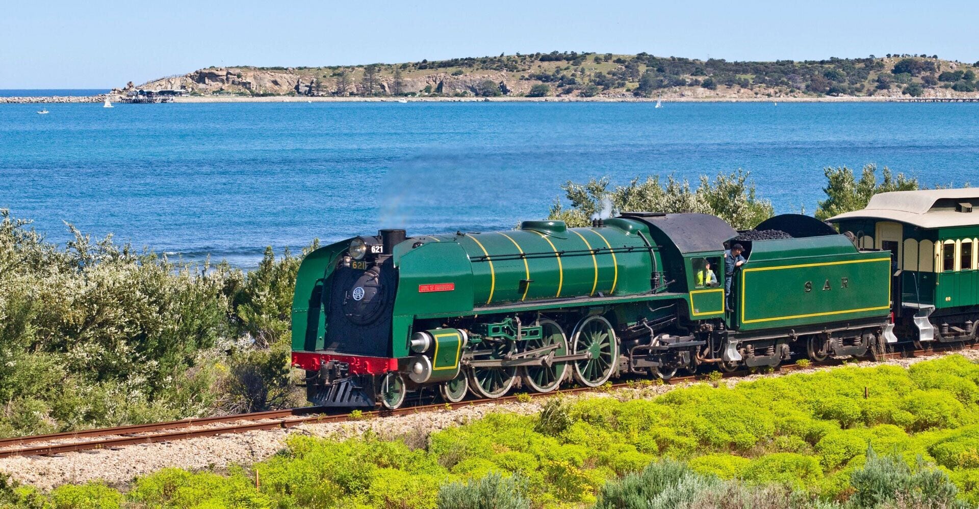 A green steam locomotive with the number 621 on the tracks, surrounded by greenery and a coastal landscape in the background.